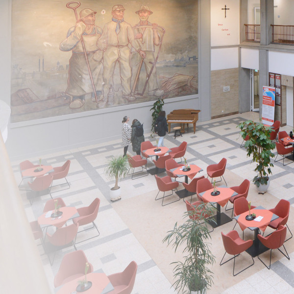 Ein großes Gebäude mit vielen Stühlen und Tischen. Es gibt eine Informationstheke und ein großes Bild an der Wand.