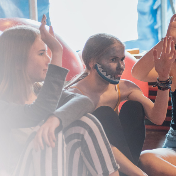 Mehrere junge Menschen sitzen zusammen. Sie haben bunte Farben im Gesicht. Sie heben die Hand und machen beim Zirkus mit.