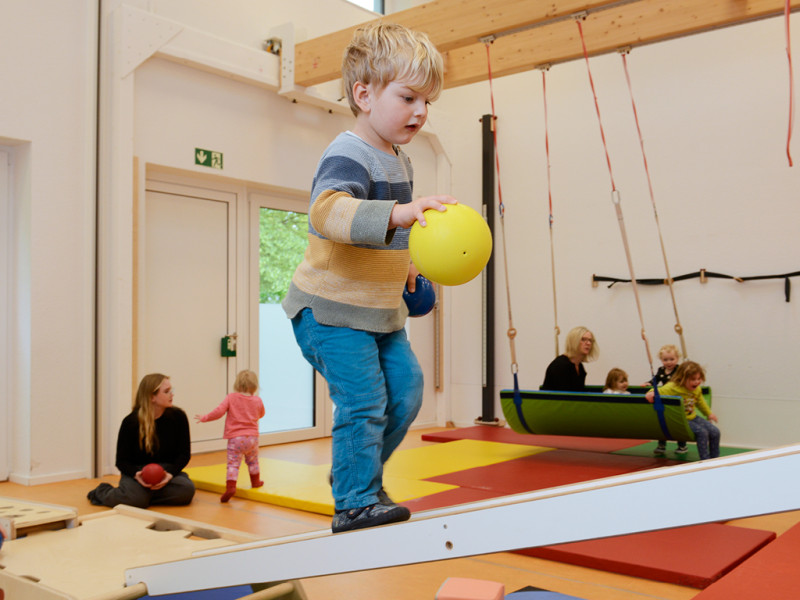 Kind balanciert mit Ball auf schräger Fläche in einem Bewegungsraum mit anderen Kindern.