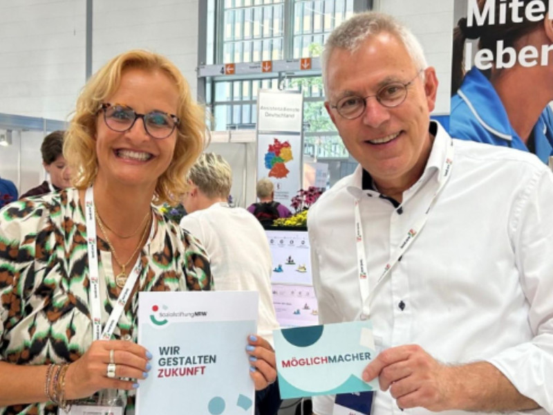 Zwei Messebesucher lächeln in die Kamera und halten Flyer mit der Aufschrift „WIR GESTALTEN ZUKUNFT“ und „MÖGLICHMACHER“ auf der REHACARE 2025.