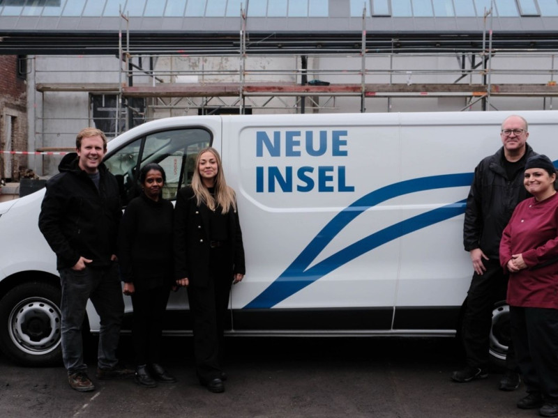 Fünf Personen stehen nebeneinander vor weißem Lieferwagen mit „Neue Insel“-Logo, Baustellenkulisse im Hintergrund