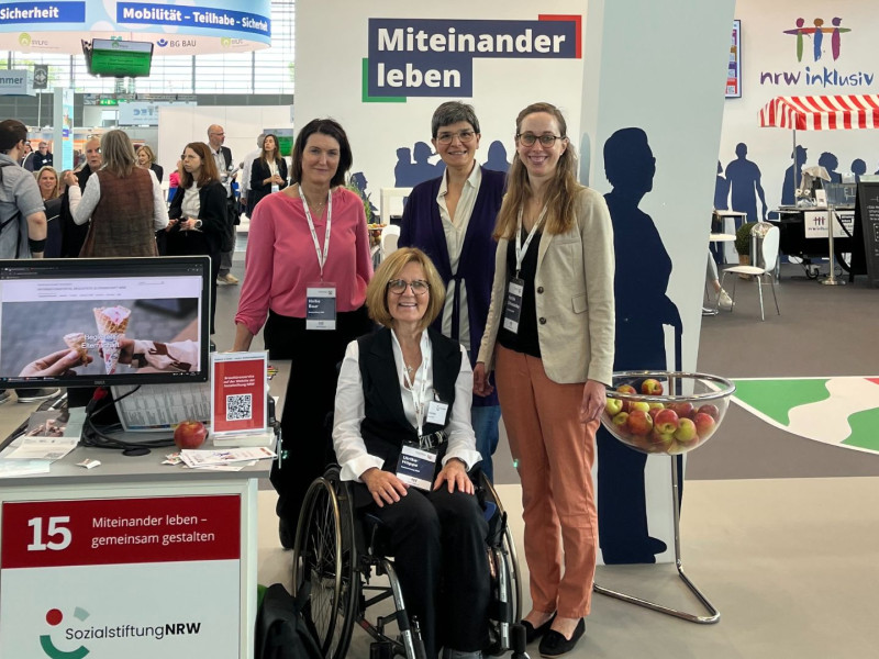 Vier Frauen, eine davon im Rollstuhl, am Stand der SozialstiftungNRW auf der REHACARE 2025 vor der Wandgrafik "Miteinander leben"