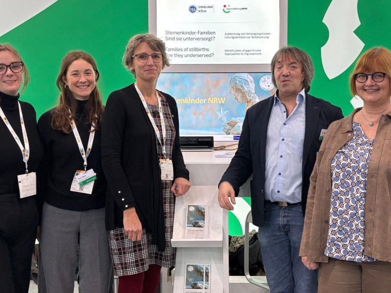 Fünf Personen stehen gemeinsam an einem Messestand des Projekts ‚Sternenkinder NRW‘. Hinter ihnen sind Informationsmaterial und ein Bildschirm mit Hinweisen zur Versorgung von Familien mit stillgeborenen Kindern zu sehen
