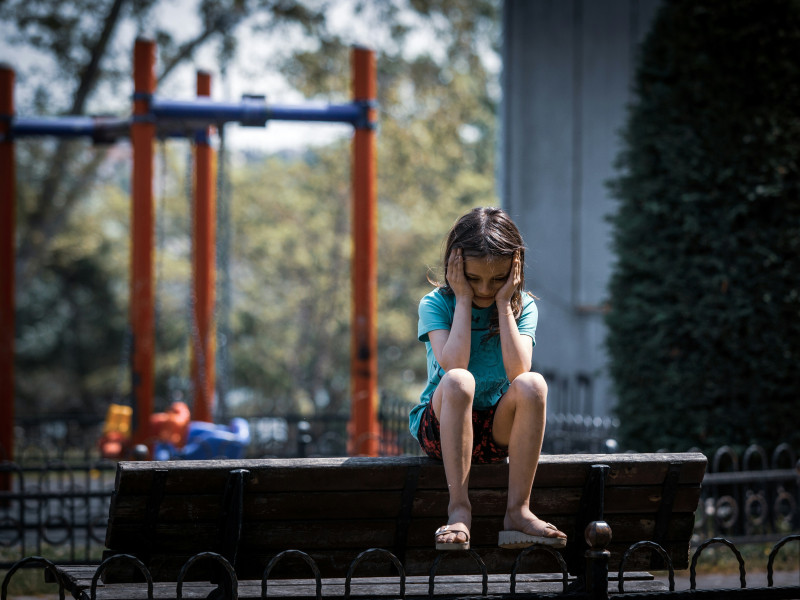 Trauriges Mädchen sitzt allein auf einer Parkbank vor einem Spielplatz und hält sich den Kopf in die Hände.