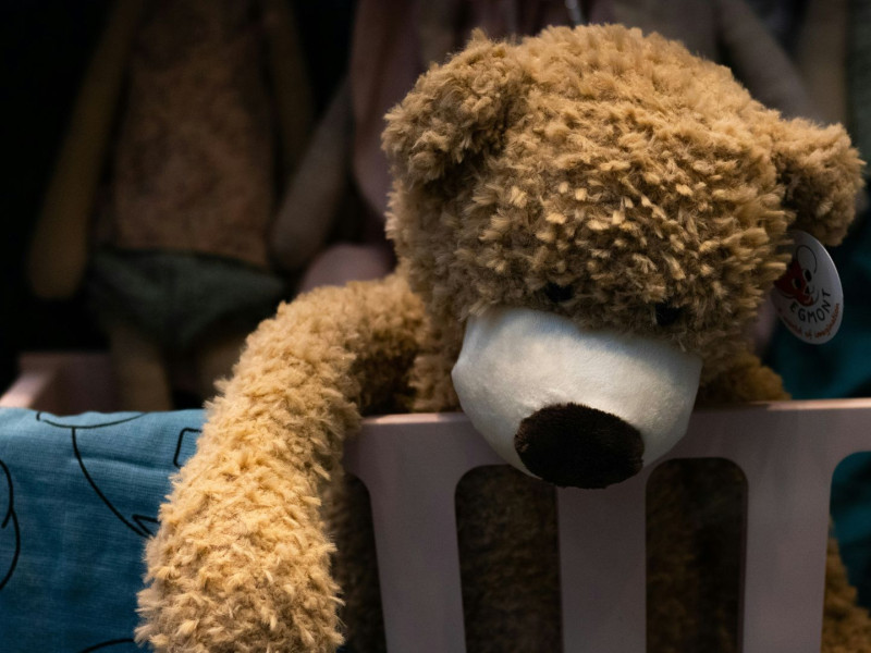 Plüsch-Teddybär liegt mit hängenden Armen über dem Rand eines Kinderbetts, im Hintergrund unscharfe Kleidung und ein Kissen mit Elefantenmotiv.