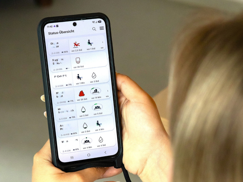 Person hält ein Smartphone mit einer Statusübersicht einer Pflege- oder Gesundheits-App in der Hand