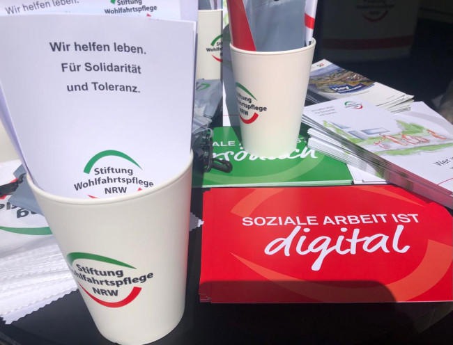 Werbematerial der Sozialstiftung NRW, hier noch Stiftung Wohlfahrtspflege. Es sind unter anderem Becher und Flyer zu sehen.