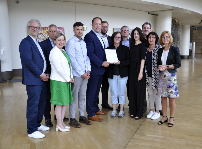 Ein Gruppenfoto, auf dem sich mehreren Personen um die überreichte wissenschaftliche Evaluation versammeln, die der Stiftungsratsvorsitzende Marco Schmitz lächelnd in die Kamera hält.