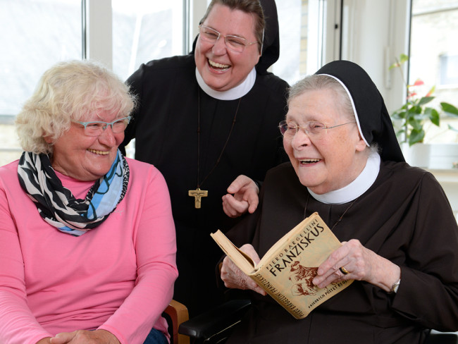 Nonne liest mit zwei Frauen ein Buch und lacht, symbolisiert Gemeinschaft.