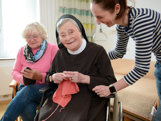 Nonne und zwei Frauen stricken und lachen zusammen, symbolisiert Gemeinschaft.