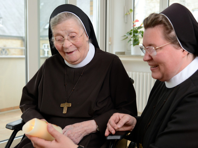 Zwei Nonnen halten eine Kerze und lächeln, symbolisiert Gemeinschaft.