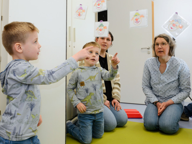 Ein Kind zeigt auf sein Spiegelbild an der Wand, während zwei Erwachsene zuschauen.