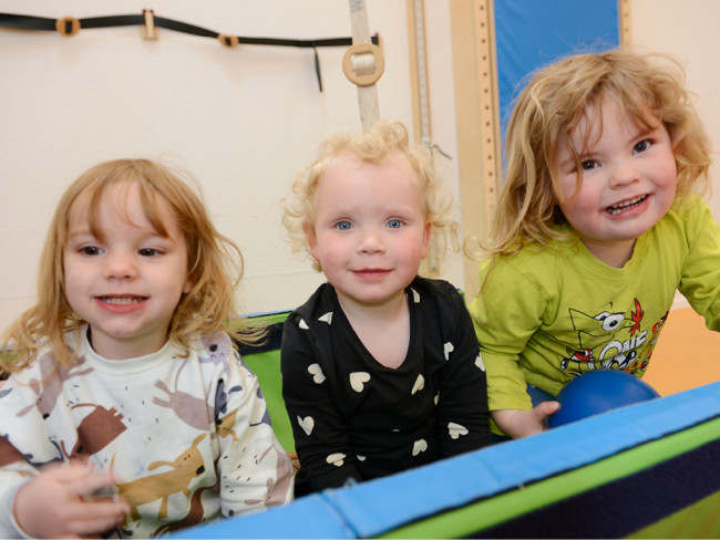 Drei lächelnde Kinder sitzen nebeneinander und schauen in die Kamera.