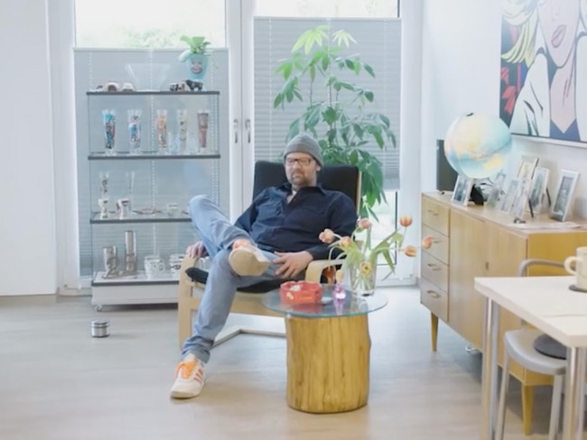 Mann sitzt entspannt in einem modernen Wohnzimmer mit Pflanzen, Kunst und Vitrinen.
