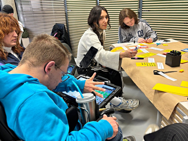 Mehrere Personen arbeiten gemeinsam an einem Tisch mit Moderationsmaterial; ein Mann im Elektrorollstuhl nutzt ein Tablet zur Kommunikation.