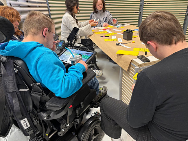 Gruppe von Personen arbeitet gemeinsam an einem Tisch mit Moderationskarten; ein Teilnehmer im Rollstuhl nutzt ein Kommunikations-Tablet.