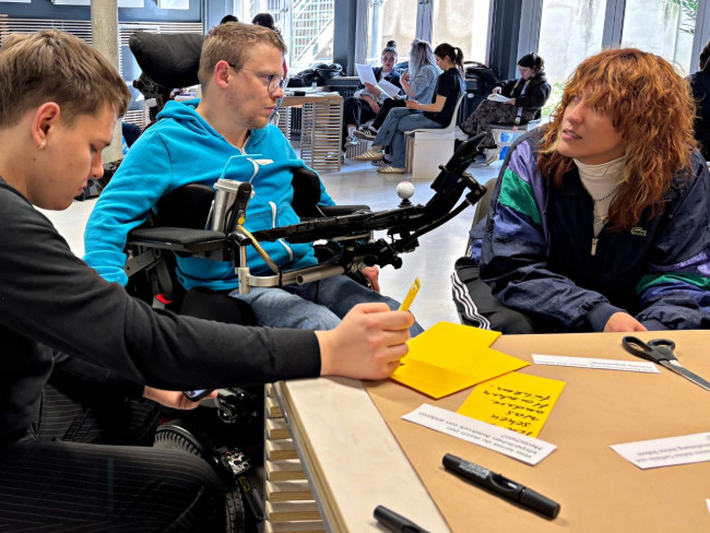 Drei Personen im Gespräch an einem Arbeitstisch mit Moderationskarten; im Hintergrund arbeiten weitere Gruppen.