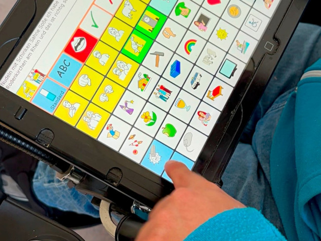 Nahaufnahme eines Tablets mit Symboltafel zur unterstützten Kommunikation; eine Hand wählt ein Symbol aus.