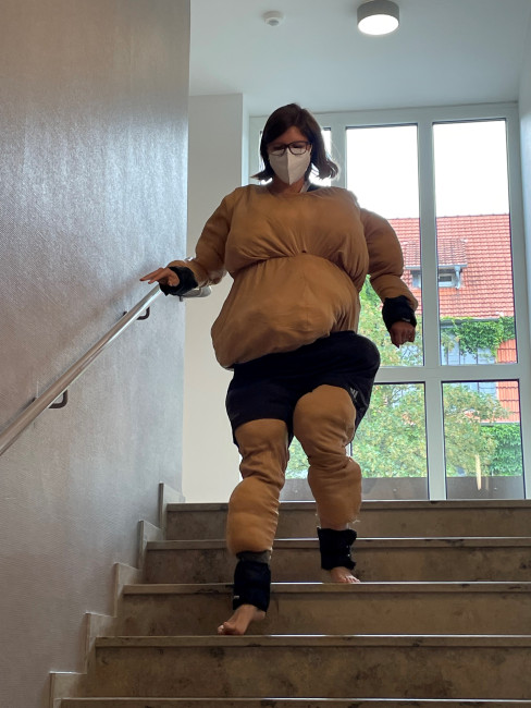 Frau im Fatsuit mit Maske steigt vorsichtig barfuß eine Treppe hinab, simuliert körperliche Einschränkungen, barrierefreies Testen in Innenraum