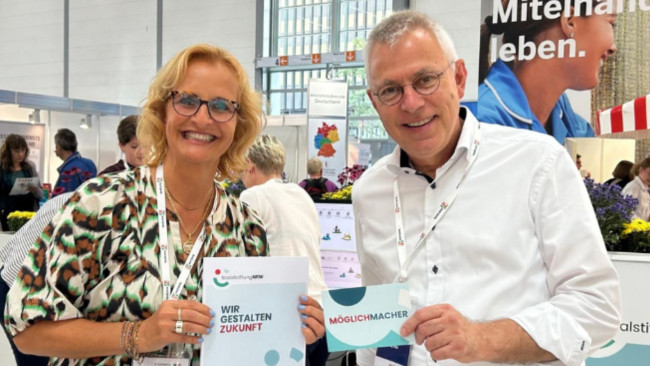 Zwei Messebesucher lächeln in die Kamera und halten Flyer mit der Aufschrift „WIR GESTALTEN ZUKUNFT“ und „MÖGLICHMACHER“ auf der REHACARE 2025.