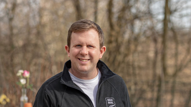 Daniel Schäfer mit kurzer Frisur und Jacke mit Logo "Neue Insel" steht draußen und lächelt freundlich in die Kamera, unscharfer Waldhintergrund.