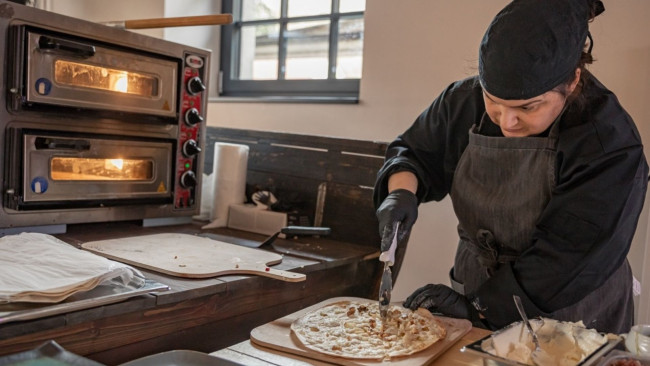 Frau in Kochkleidung bereitet konzentriert Flammkuchen in moderner Küche vor, daneben ein Doppelofen mit eingeschaltetem Licht