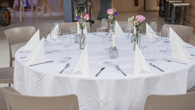 Runde Tafel festlich gedeckt mit weißen Servietten, Gläsern, Besteck und Blumenvasen in heller Umgebung mit moderner Einrichtung