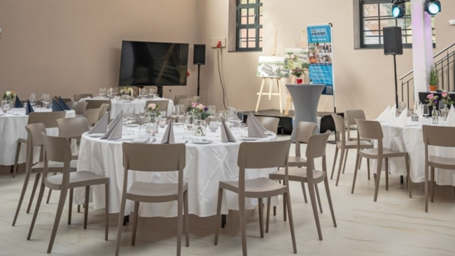 Mehrere runde Tische mit weißen Tischdecken, Servietten und Blumendekoration in hellem, modernem Saal mit Leinwand und Stehtischen