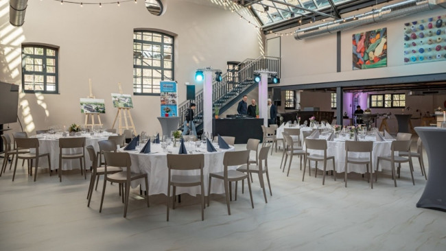Großer Saal mit runden Tischen, Lichterkette unter Glasdach, DJ-Pult und Wandkunst; freundliche, helle Eventatmosphäre