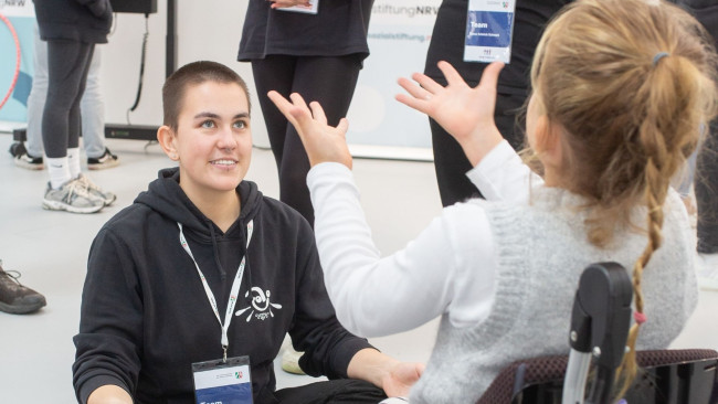 Junge Frau und Kind im Rollstuhl interagieren spielerisch auf der REHACARE; beide lachen und sind Teil eines inklusiven Mitmachangebots am Messestand.