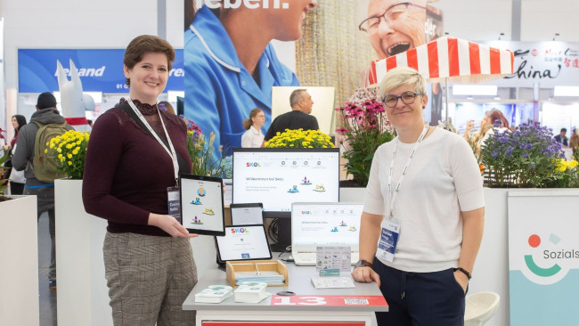 Zwei Personen präsentieren digitale Tools und Infomaterial zur SKoL-Toolbox für selbstbestimmtes Leben im Alltag auf der REHACARE 2025.