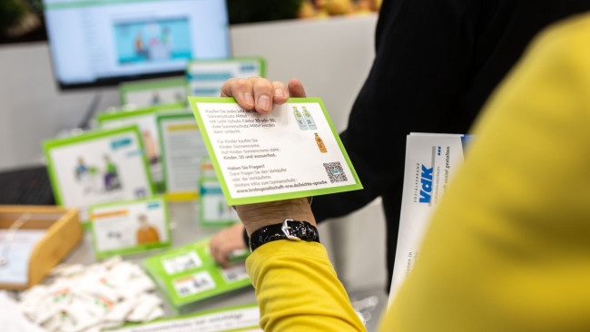 Hand hält Flyer mit Tipps zum Sonnenschutz in leichter Sprache, Informationsstand im Hintergrund, Fokus auf verständliche Gesundheitskommunikation.