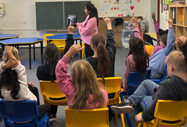 Kinder schauen zur Lehrerin, die ein Tablet hochhält und melden sich.