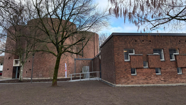 Außenansicht der ehemaligen Kirche St. Peter Morus in Leverkusen mit rotem Backsteinbau, rundem Kirchenschiff und modernem Anbau, umgeben von Bäumen.