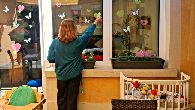 Person klebt bunte Papierherzen und Schmetterlinge an ein Fenster in einem Innenhof.