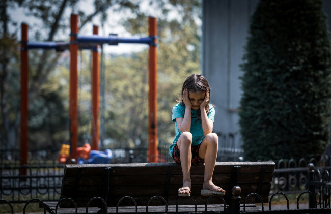 Trauriges Mädchen sitzt allein auf einer Parkbank vor einem Spielplatz und hält sich den Kopf in die Hände.