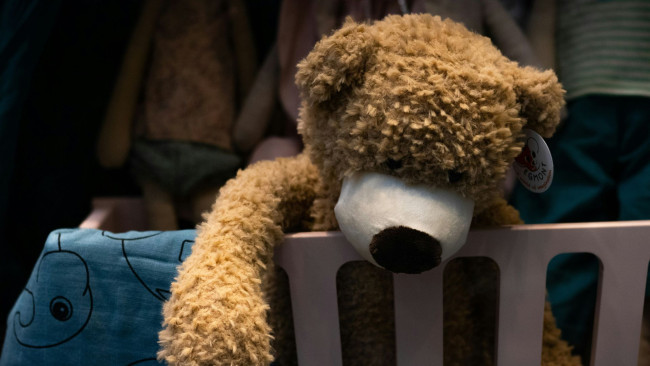 Plüsch-Teddybär liegt mit hängenden Armen über dem Rand eines Kinderbetts, im Hintergrund unscharfe Kleidung und ein Kissen mit Elefantenmotiv.