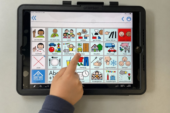Bildschirmansicht eines Talkers mit Symbolen für verschiedene Kontexte. Kinderhand zeigt auf "Haus"