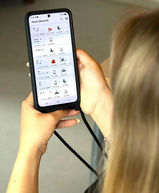 Person hält ein Smartphone mit einer Statusübersicht einer Pflege- oder Gesundheits-App in der Hand