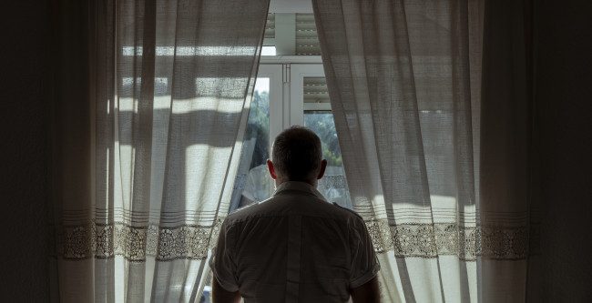Mann steht zwischen Vorhängen an einem Fenster und blickt nach draußen.