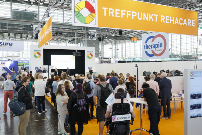 Treffpunkt REHACARE 2025 auf der Messe Düsseldorf mit zahlreichen Besucherinnen und Besuchern vor der Veranstaltungsbühne.