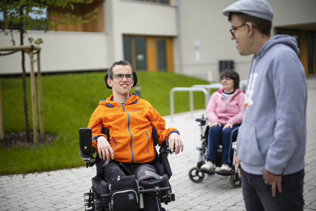 Drei junge Erwachsene, darunter zwei Rollstuhlnutzer, unterhalten sich im Außenbereich.